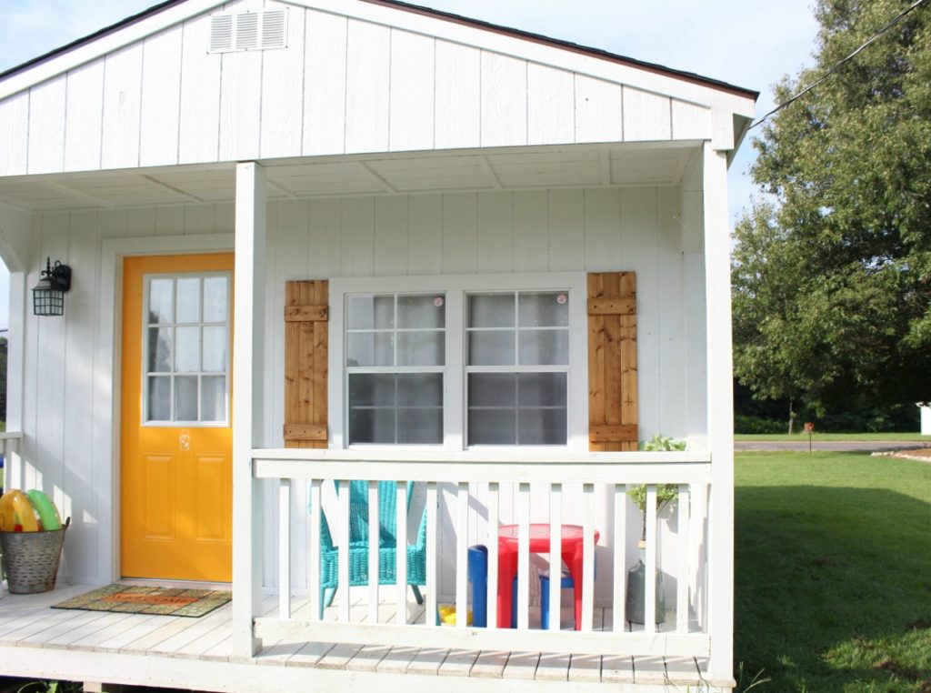 Front of beautiful white she shed with natural wooden shutters and yellow front door