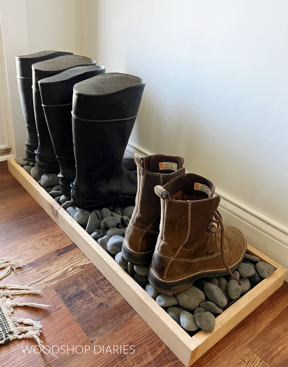 Wooden boot tray with black decorative rocks inside next to front door--two pair of rainboots and a pair of work boots on top