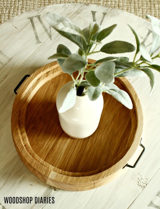 round serving tray sitting on coffee table with vase of faux greenery sitting on it.