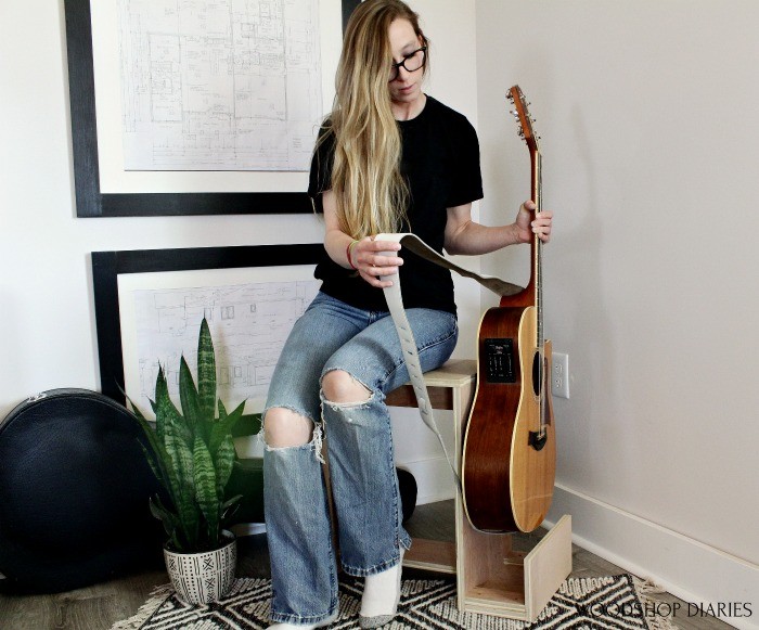 Shara Woodshop Diaries sitting on plywood stool picking up guitar out of stand to play