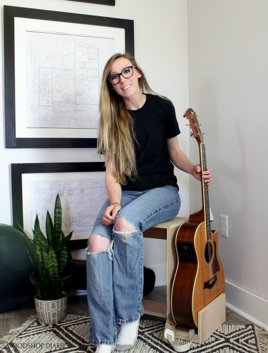 Shara sitting on scrap wood stool holding guitar in stand