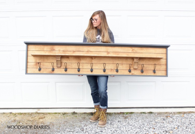 Shara in front of garage door holding long DIY coat rack with shelf--wood back panel with black frame around outside