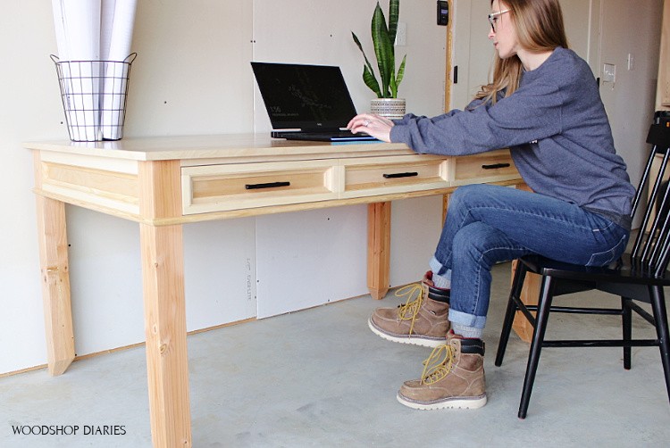 Shara Woodshop Diaries sitting at large writing desk on laptop in black chair