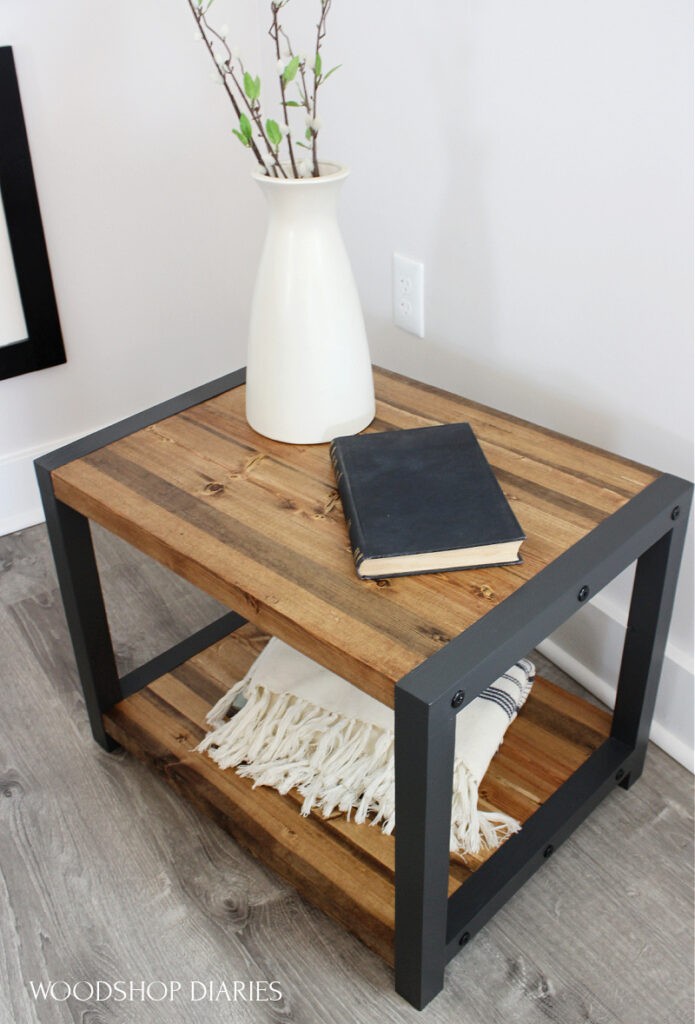 Angled view of DIY black and wood end table with timber screws on sides. Black frames and wood top and bottom shelves with vase and book on top