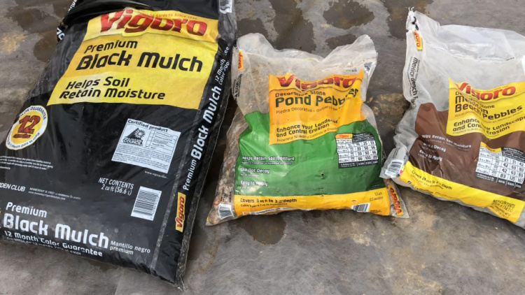 Black mulch, pond pebbles, and Mexican beach pebble bags laying side by side on ground