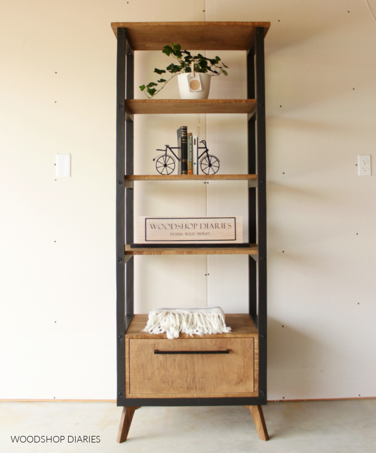 Black and wood mid century modern industrial style open bookshelf with drawer at bottom.  Styled with plants, books, and blanket