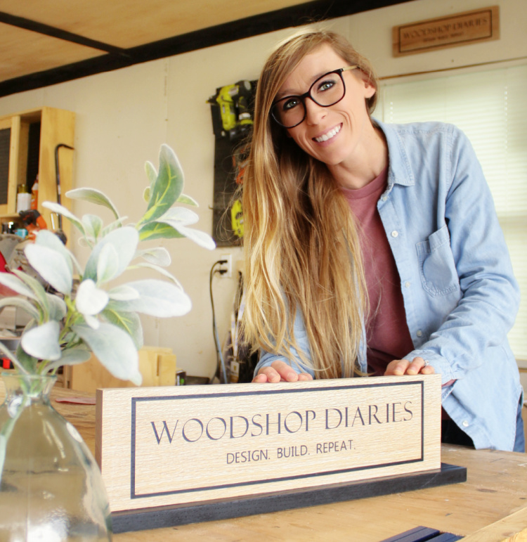 Shara Woodshop Diaries with DIY custom wooden signs in workshop--one on wall and one on base plate sitting on workbench