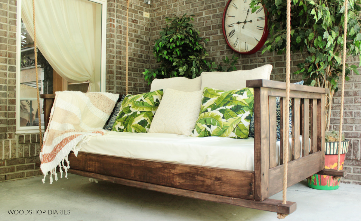 Completed DIY porch swing bed hanging on brick porch with ropes on each corner and green and navy pillows along back side of swing.