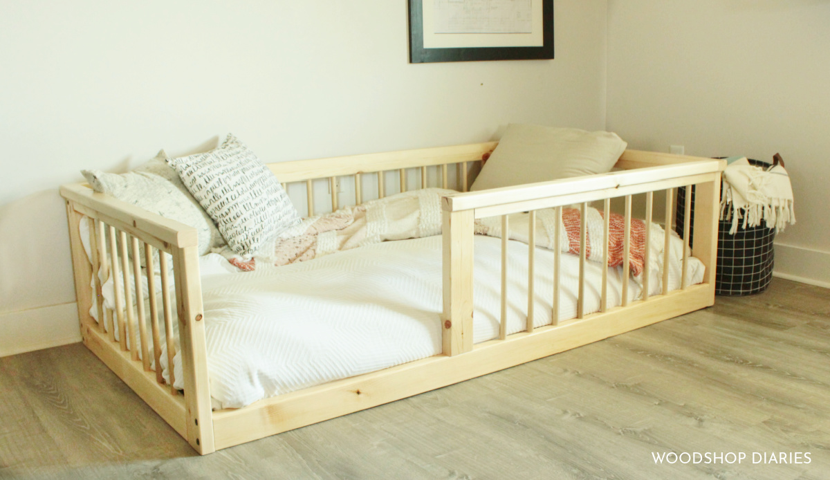 Simple pine wood twin size floor bed frame with side rails.  White comforter and burnt orange bedding against a light great wall