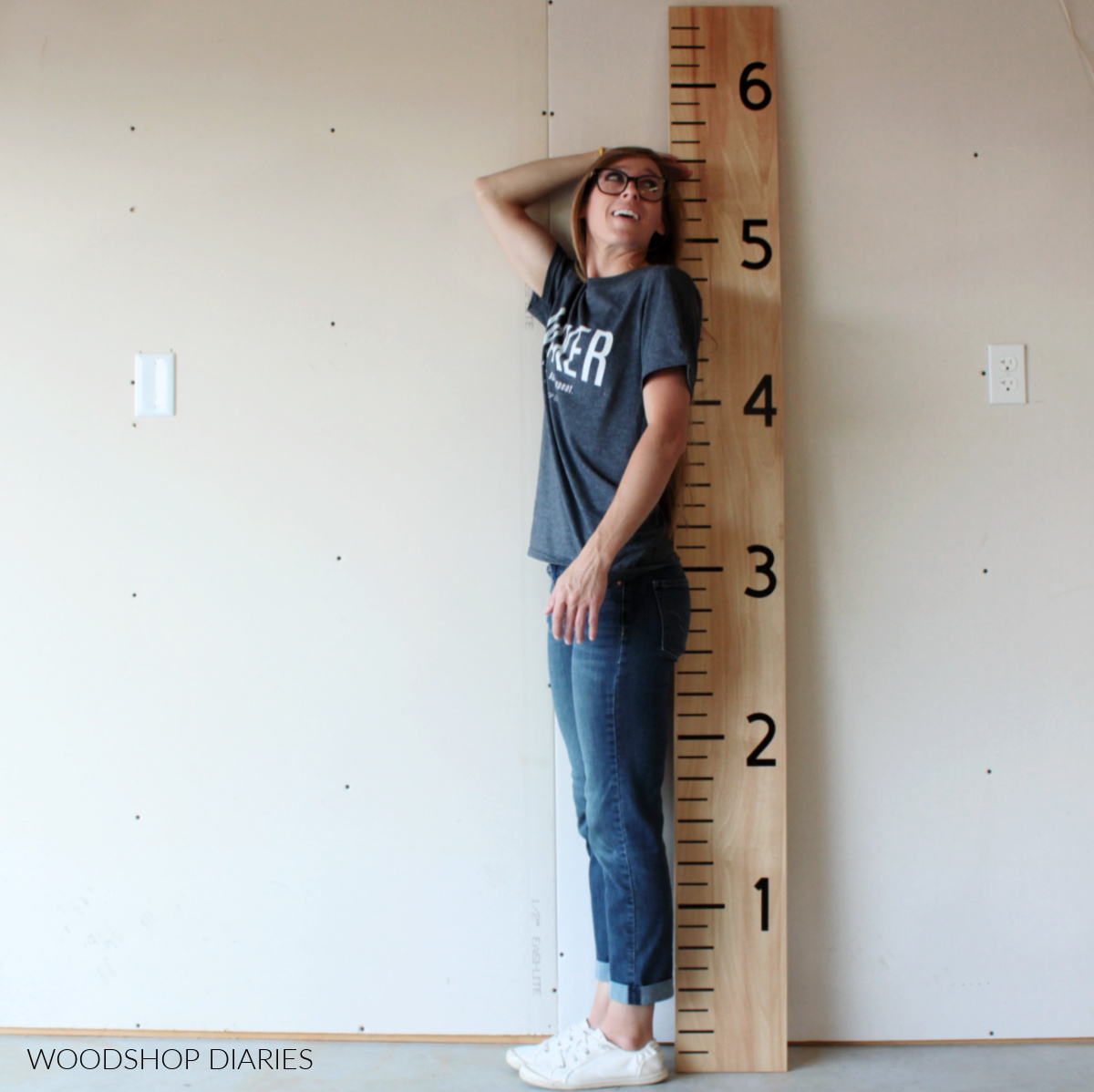 Shara Woodshop Diaries standing next to life size giant ruler board growth chart checking her height