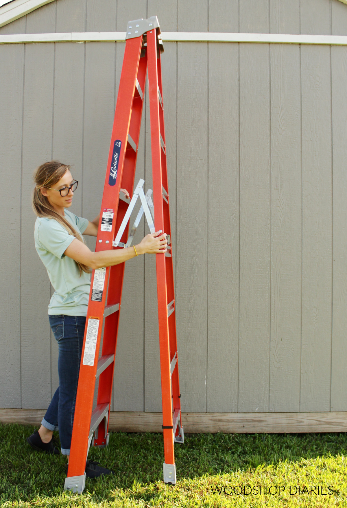 Shara Woodshop Diaries folding up 8 ft step ladder next to shed