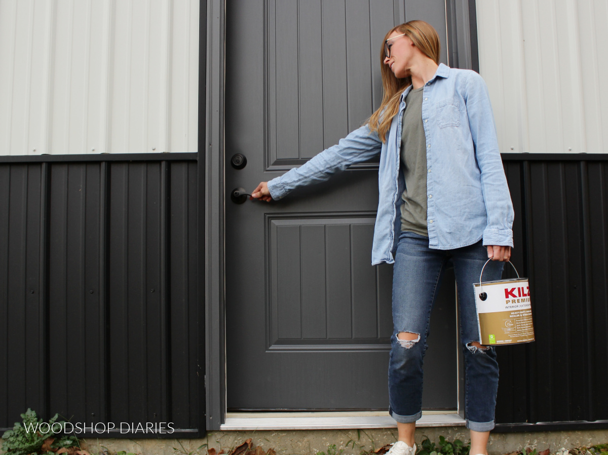 Shara Woodshop Diaries holding a can of primer and reaching to open freshly painted grey door against black and white metal building