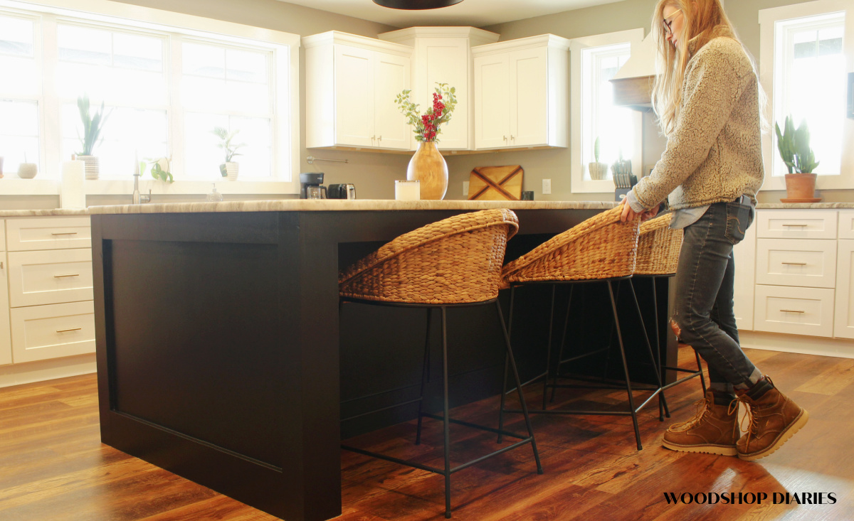 Shara pushing wicker barstool up underneath black kitchen island in white cabinet kitchen