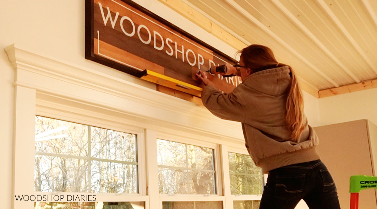 Shara Woodshop Diaries hanging large wooden sign in workshop above windows