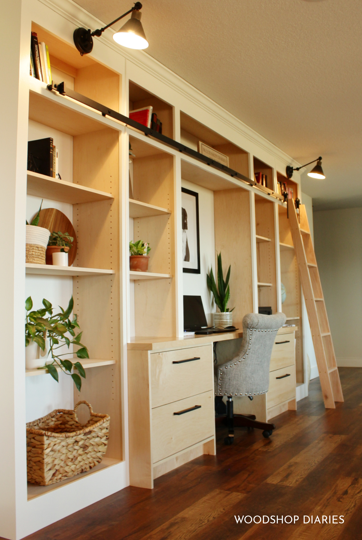 White and wood bookshelf unit with desk in the middle with rolling ladder and black sconce lights and grey office chair