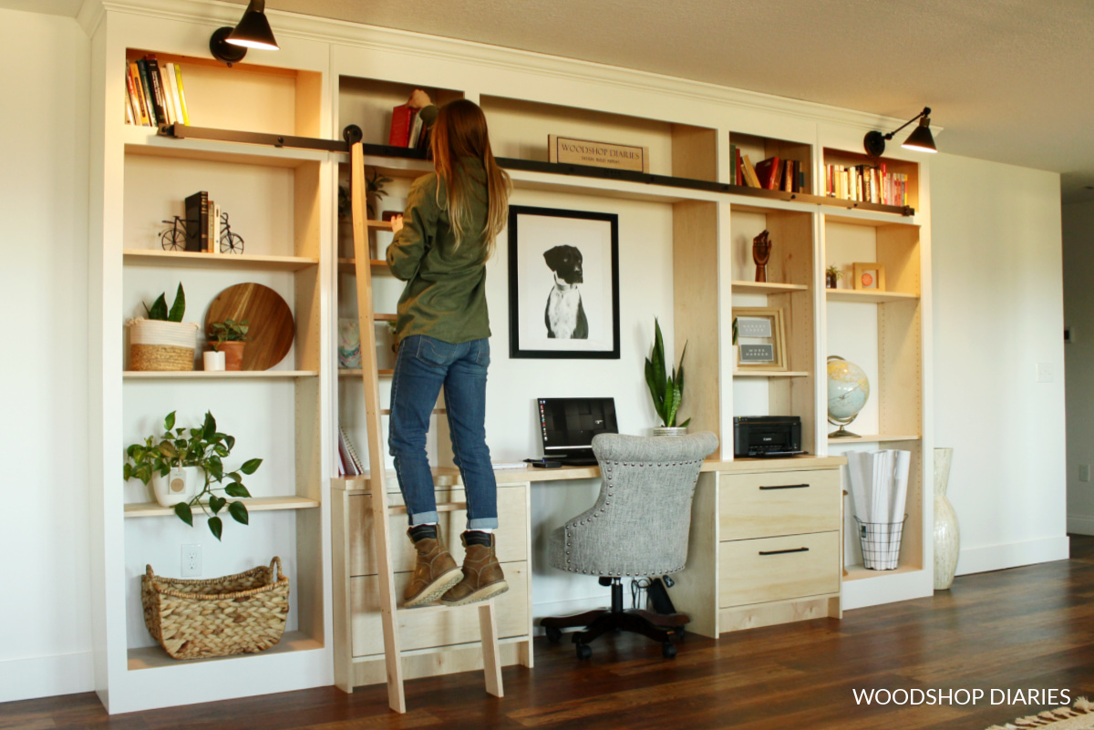 Shara Woodshop Diaries on rolling library ladder picking a book off the shelf