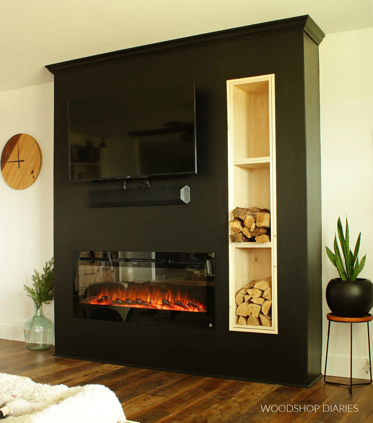 White walls with black DIY electric fireplace wall with TV. Stacked wood cubbies on left side with firewood