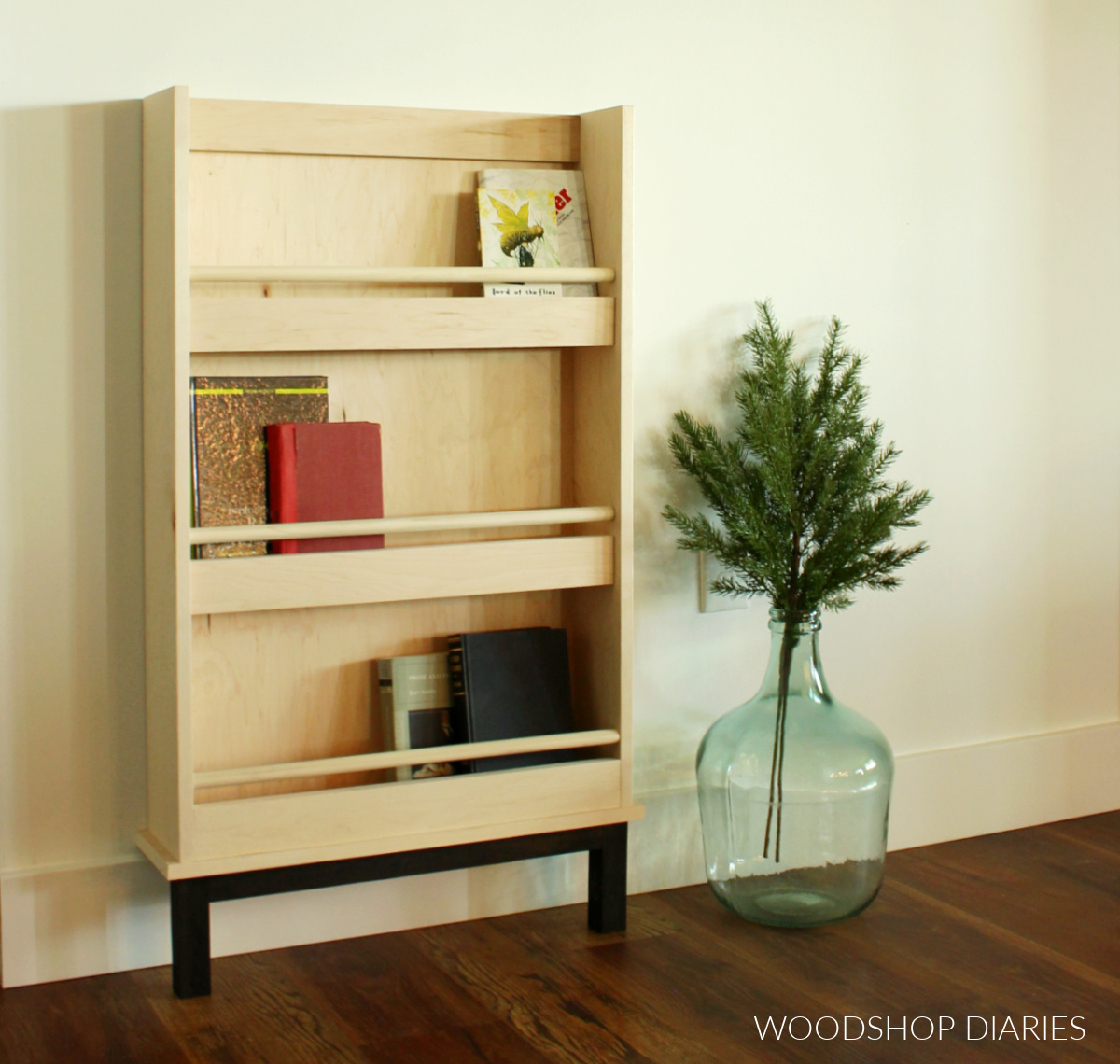 Plywood book shelf rack with various books inside leaning against wall next to glass vase