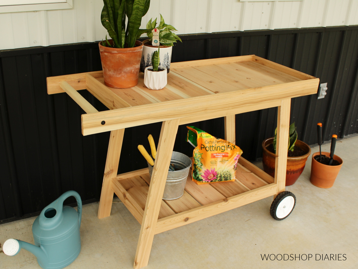 Completed cedar potting bench with handle and wheels against black and white siding with plants around