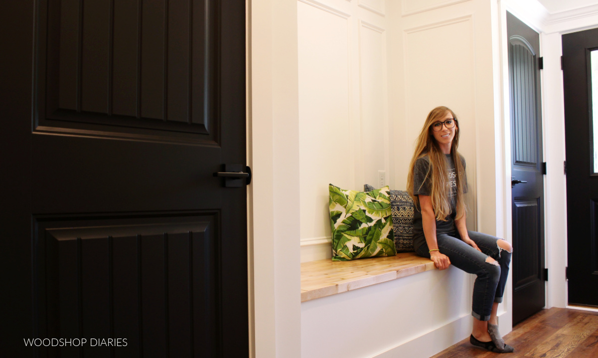 Shara Woodshop Diaries sitting on built in bench in hallway with white walls and wall trim and black doors with wooden bench seat top--Hallway makeover "after" image