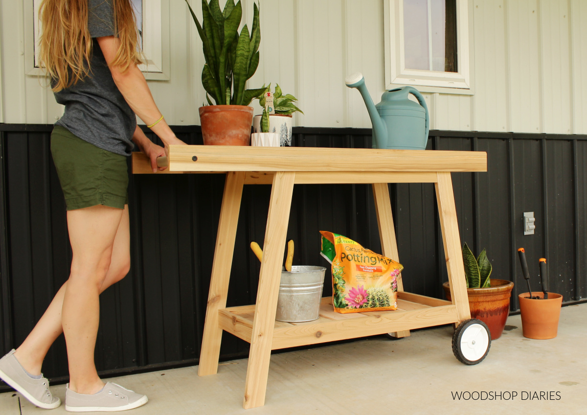 Shara Woodshop Diaries pushing DIY potting bench cart with wheels on porch