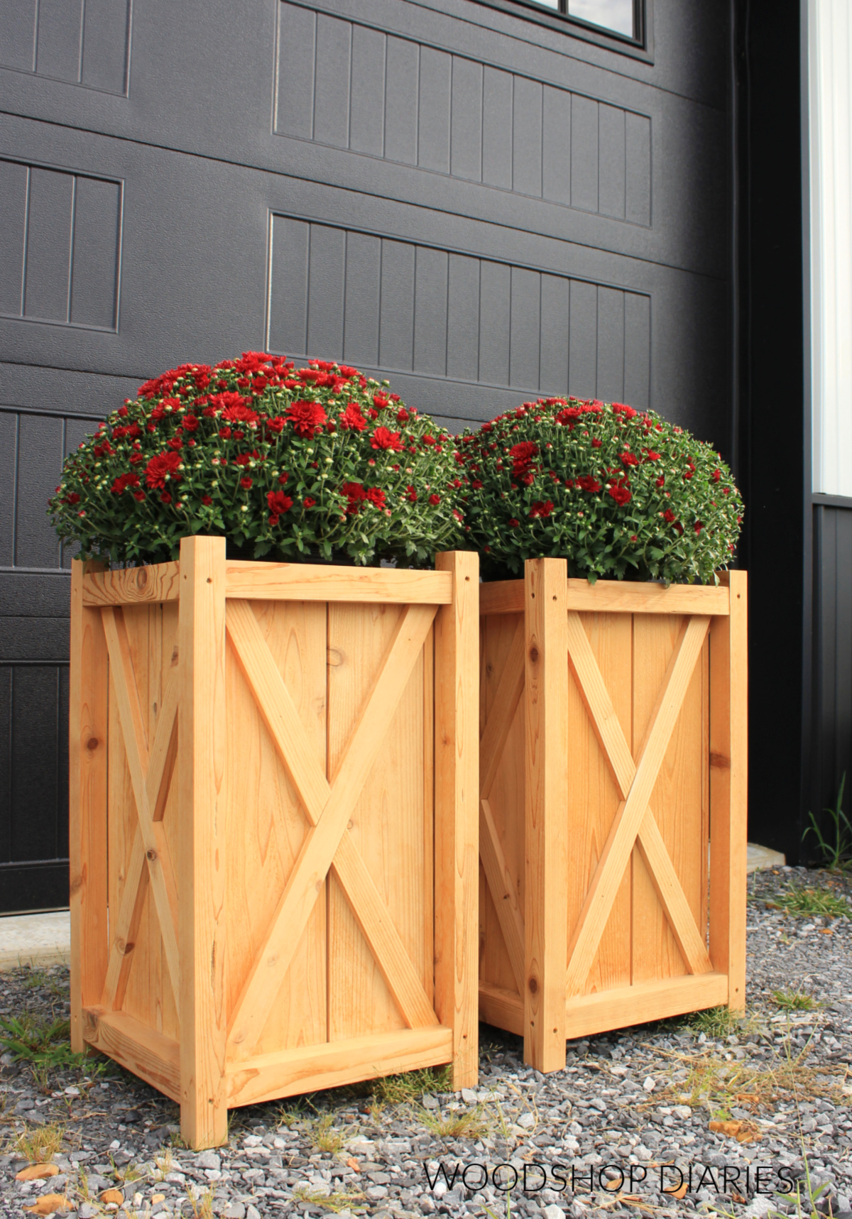 Pair of completed DIY wooden planters with X trim detail in front of black garage door with red mums inside