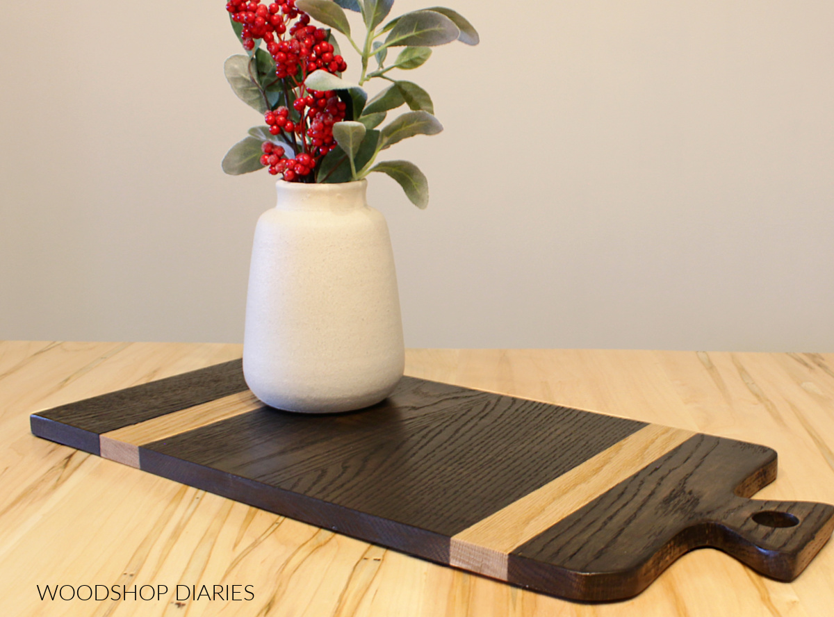 Stained black oak and natural oak stripes DIY cutting board laid out on table top with vase on top