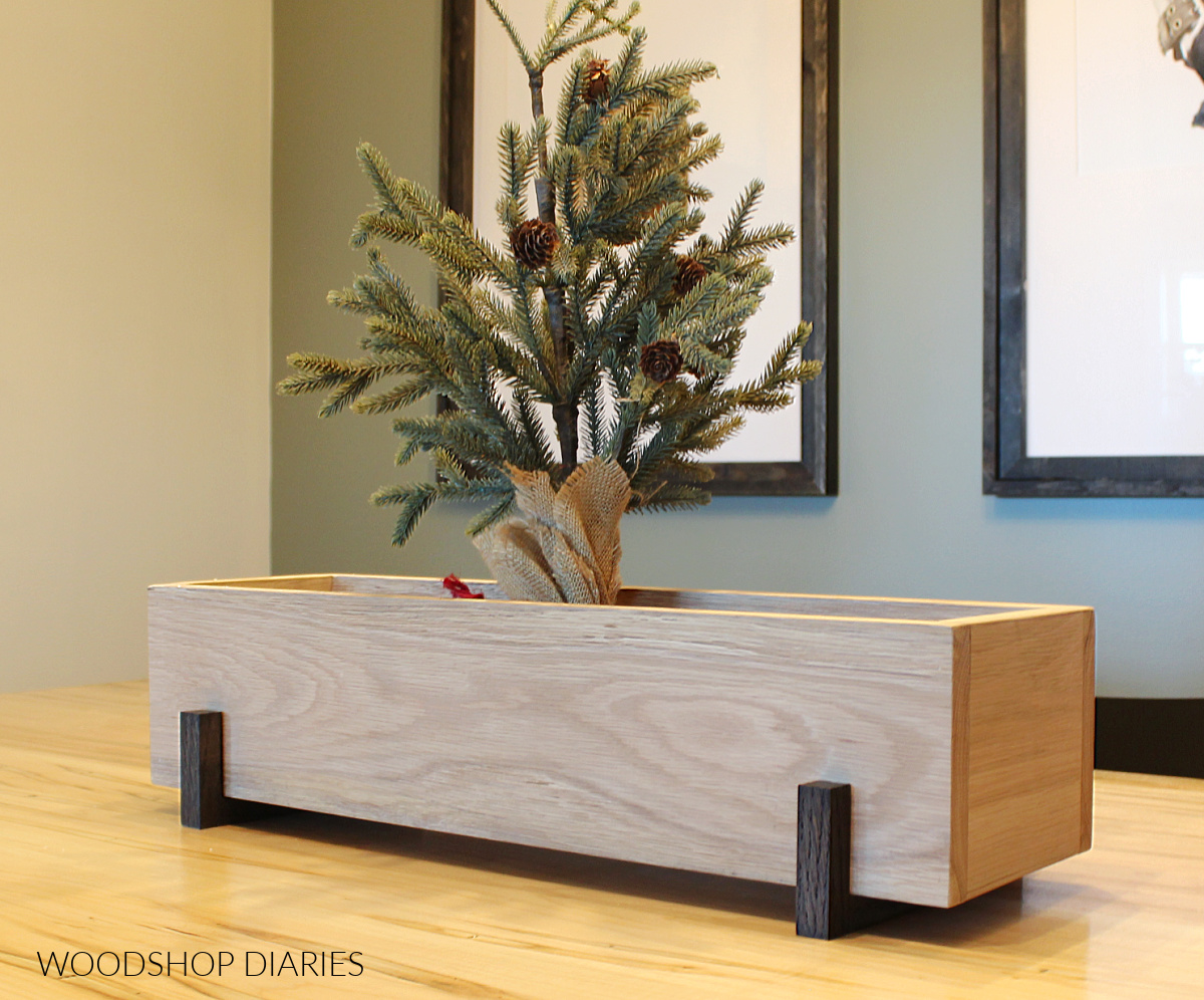 White oak centerpiece box with modern black risers on dining table with mini Christmas tree inside