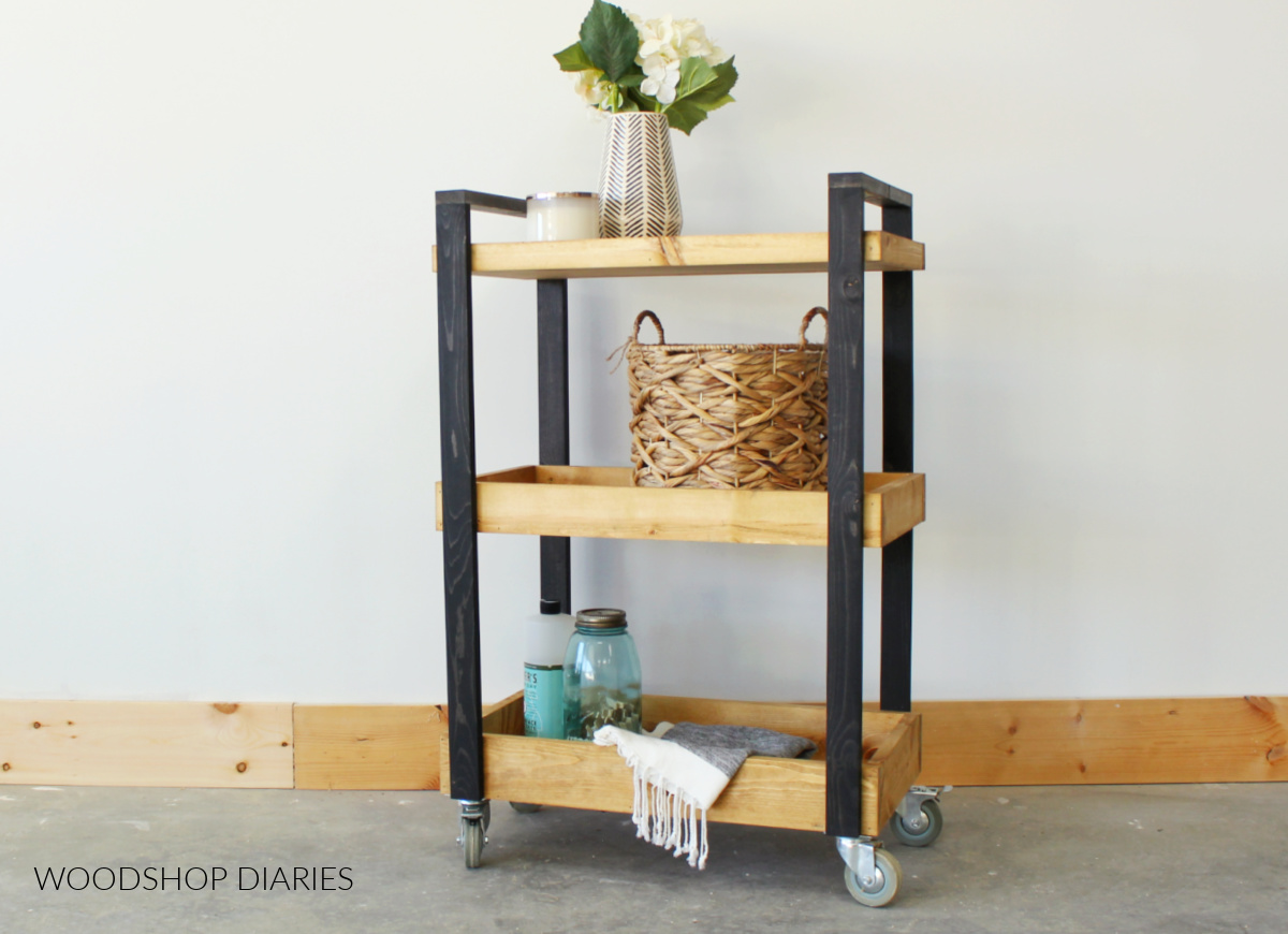 Black and wood tone three tiered mobile storage cart with cleaning supplies and storage basket 