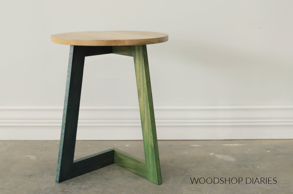 Simple modern C table with natural wood top and blue and green stained table base against white wall