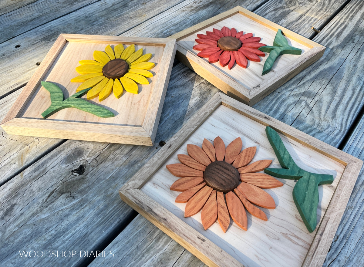 Three different color wooden sunflower art pieces laid against weathered wood backdrop--one yellow, one orange, one red flower with green stem