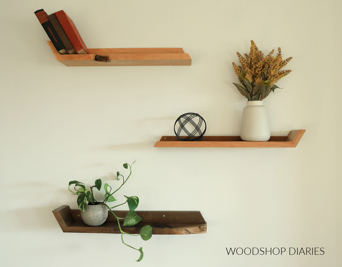 Three different colored wooded angled DIY floating shelves on white wall with books, vase, and plant sitting on them