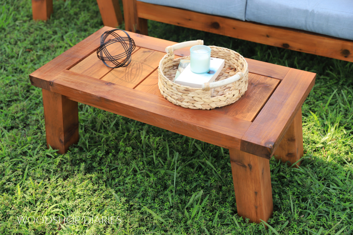 Completed DIY outdoor coffee table sitting in grass in front of loveseat with wicker basket and candle on top