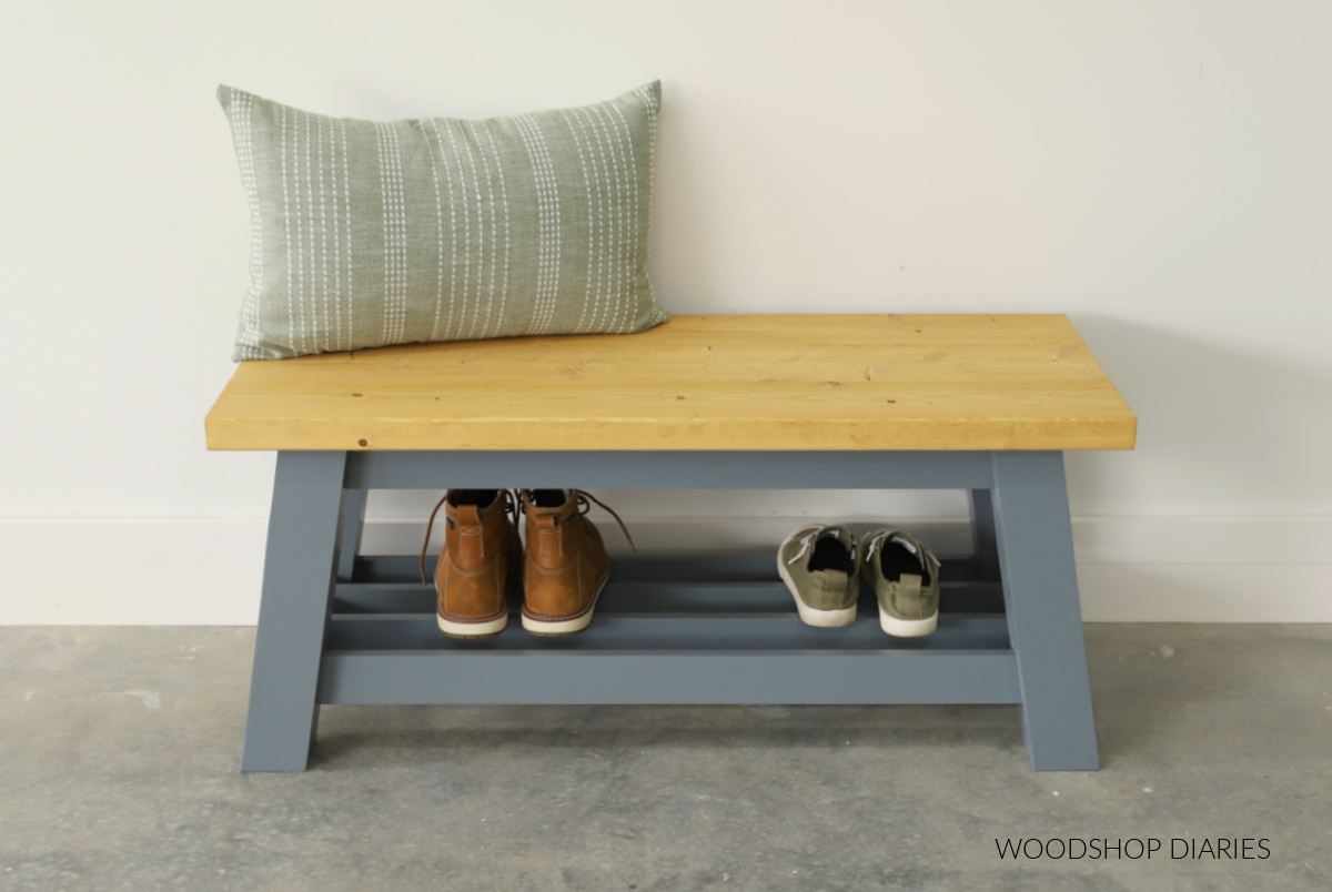 Blue gray wooden DIY shoe bench with boots and sneakers on bottom shelf and green pillow on bench seat.