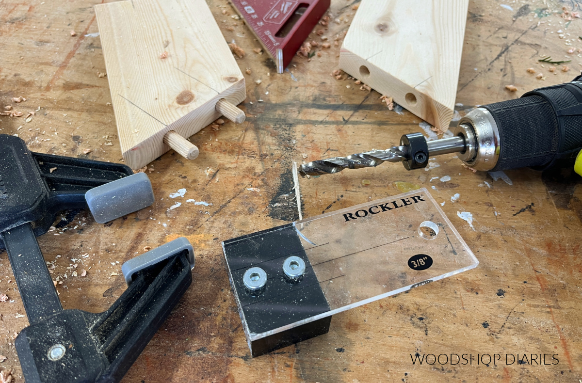 Dowel jig, drill, clamp, and square laying on workbench with two boards with dowel holes drilled out and dowel pins inserted
