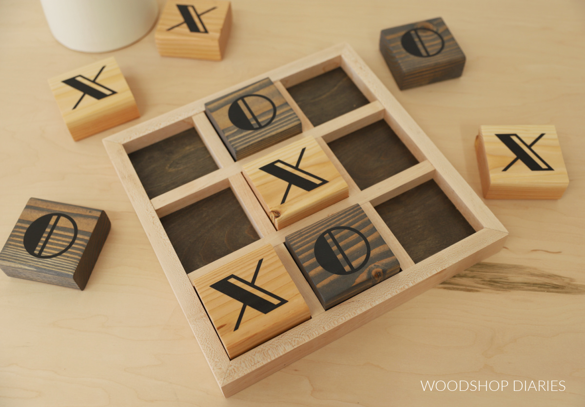 Black and wood two tone wooden tic tac toe game board on workbench with game pieces spread out around it