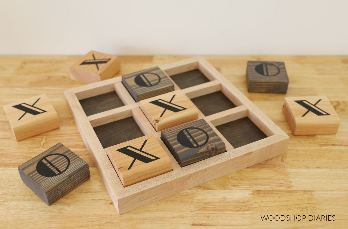 Wooden tic tac toe board with contrasting colors--black and gray stain with natural wood tones. Xs and Os game pieces spread out around the board