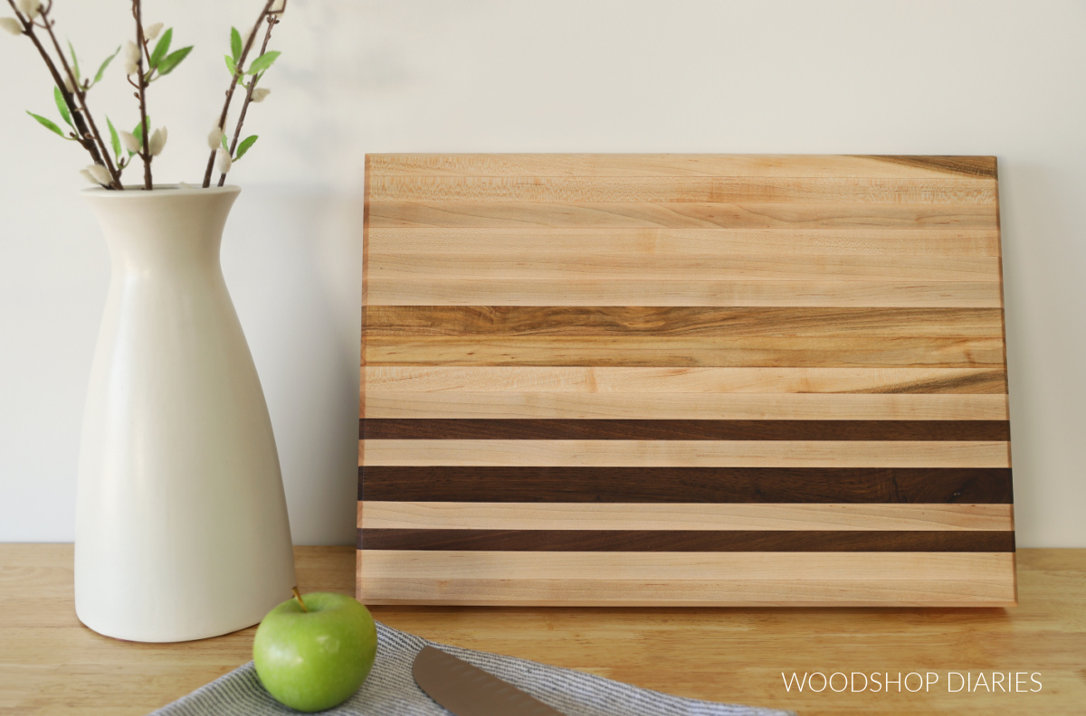 completed maple and walnut cutting board leaning against the wall next to white vase with green apple, knife and towel on countertop