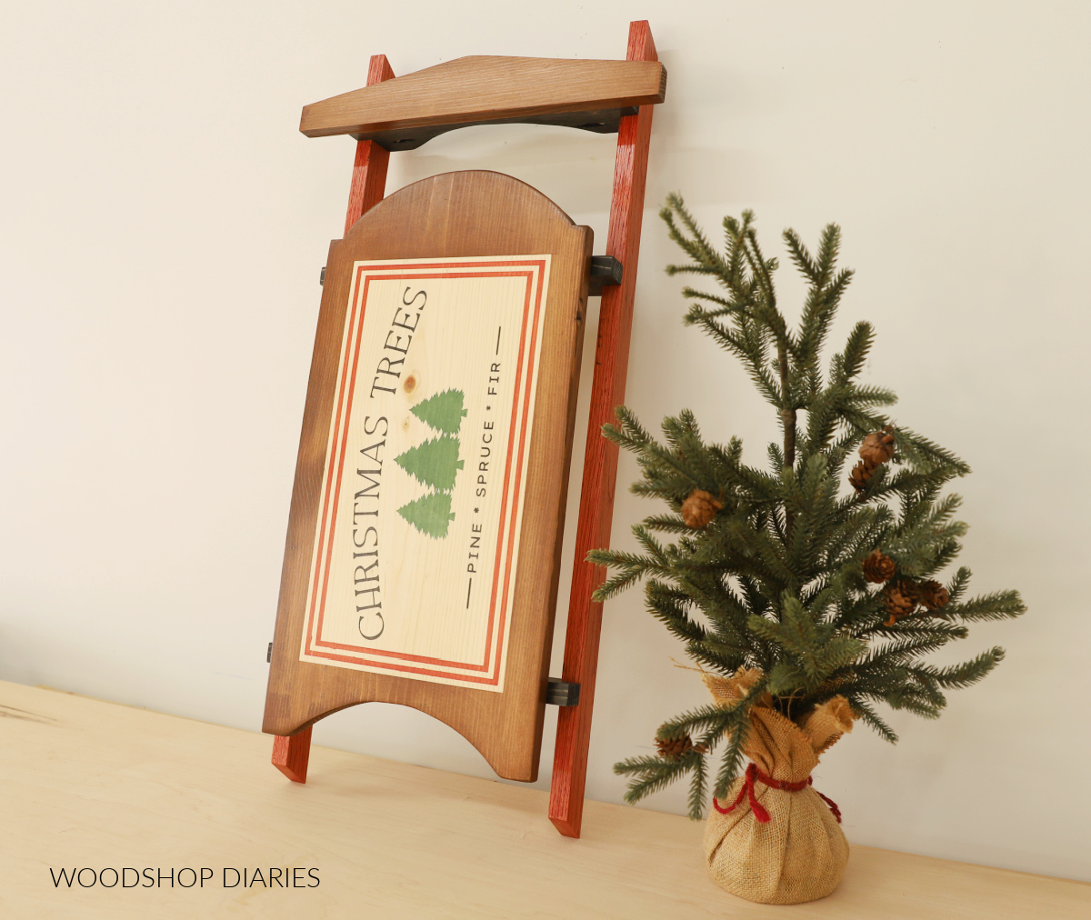 DIY Wooden Sled propped up against wall next to faux Christmas tree. Sled seat has stenciled design showing Christmas Trees