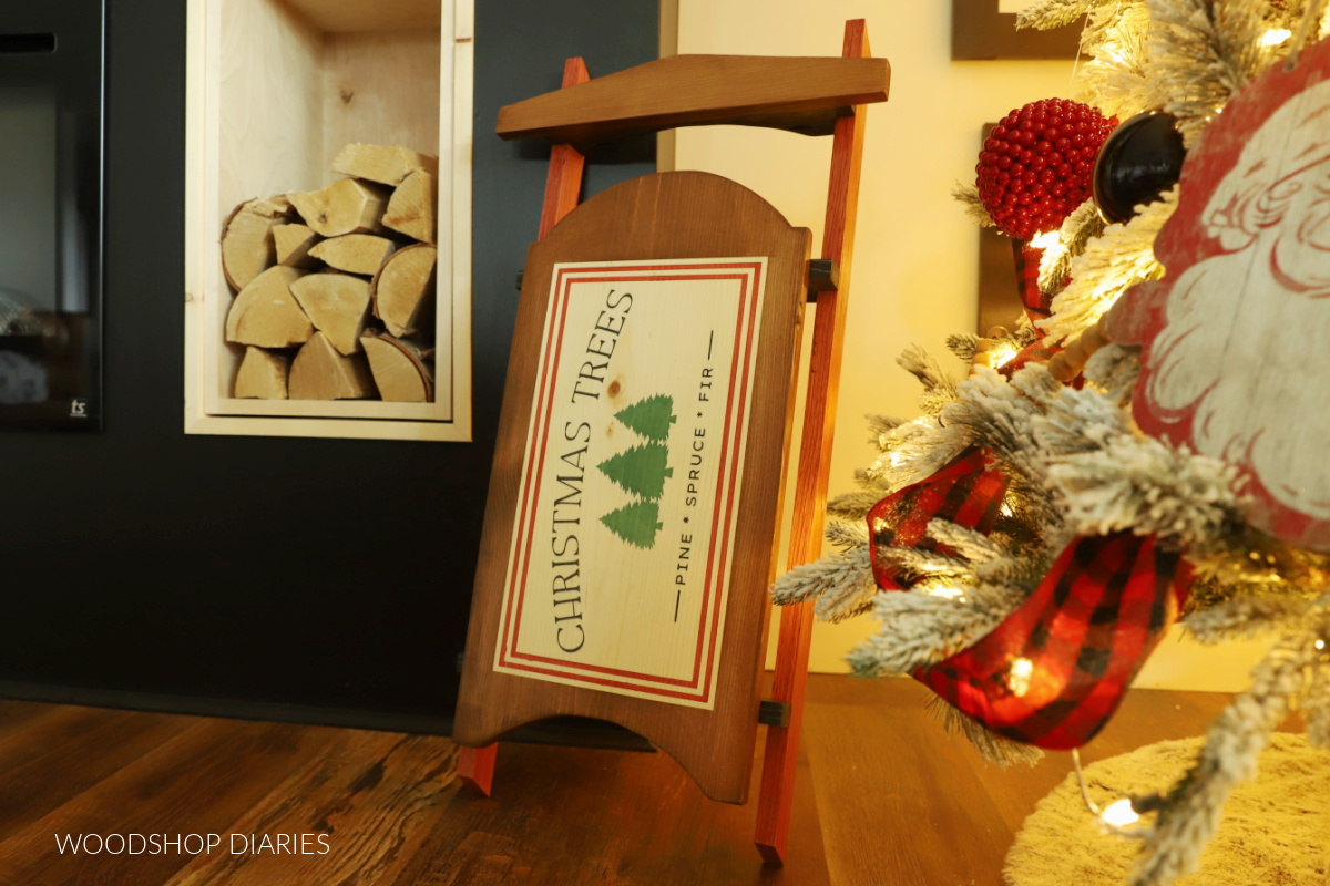 Decorative wooden sled propped up against fireplace next to Christmas tree with stenciled design that says "Christmas Trees Pine Spruce Fir"