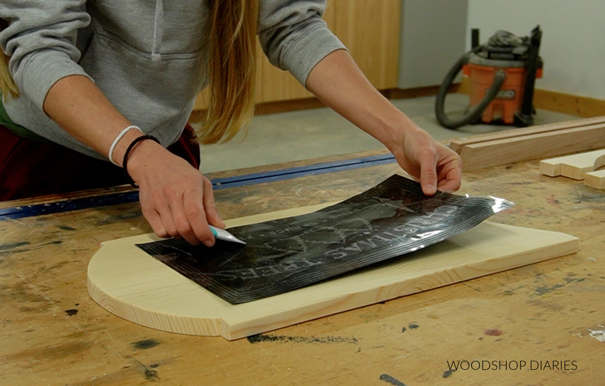 Shara Woodshop Diaries applying vinyl stencil to wooden sled seat board on workbench