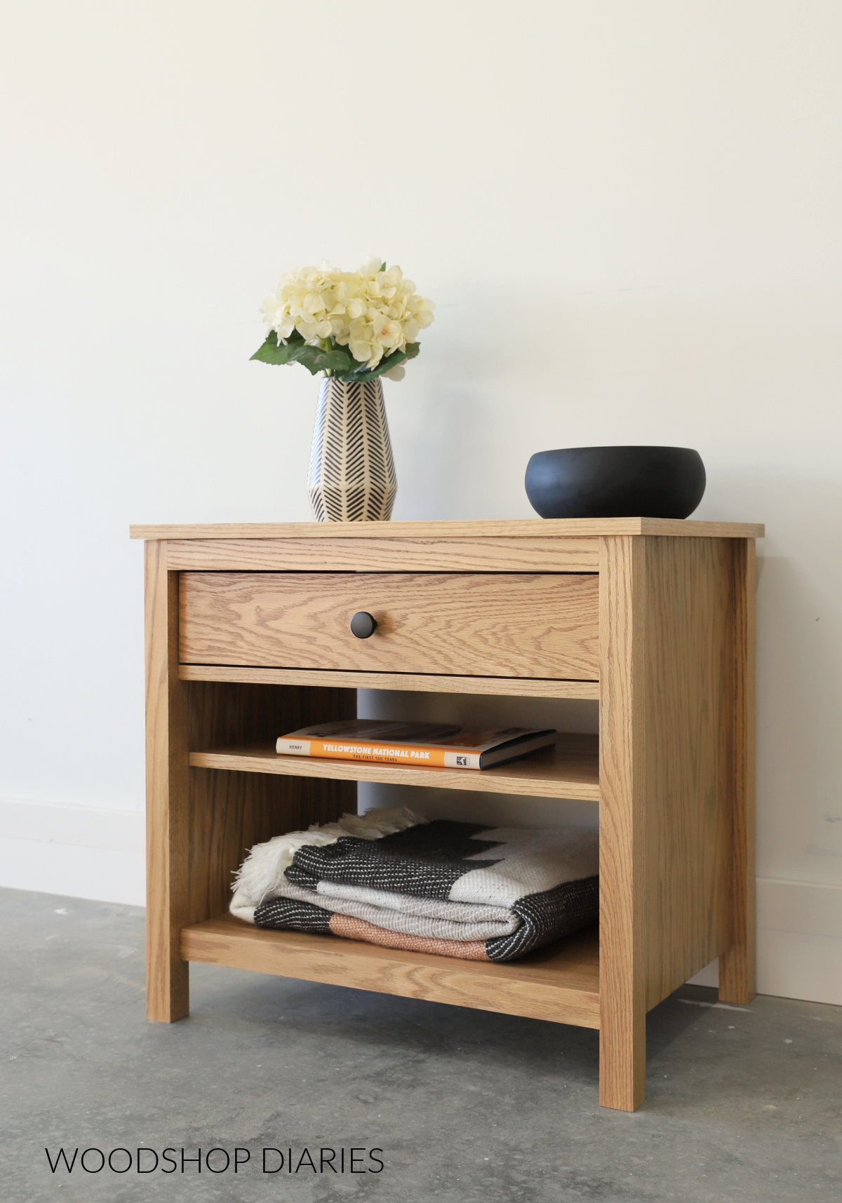 Red Oak Modern Nightstand with drawer and two shelves against white wall with vase on top and books, blankets on shelves