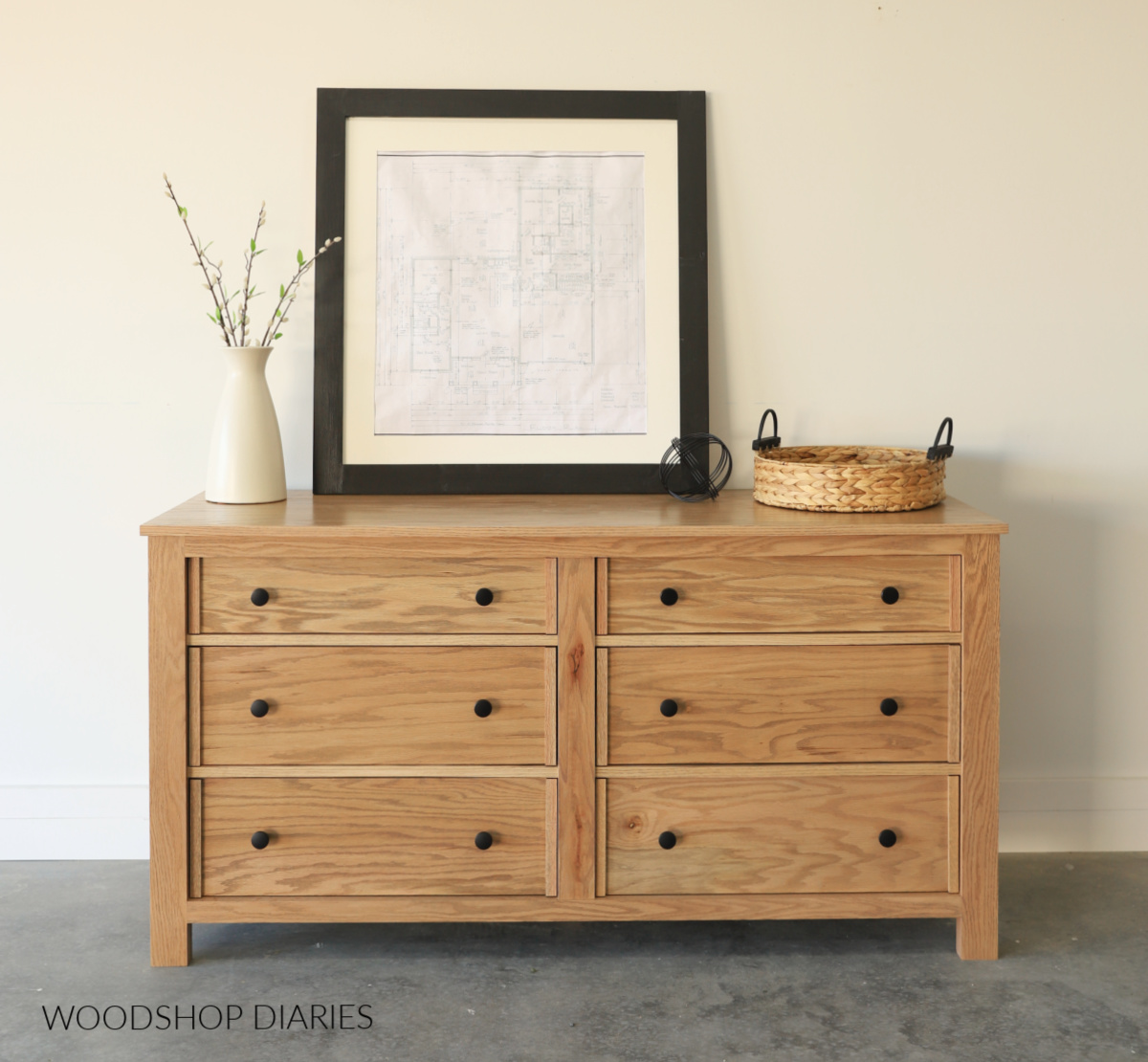 Red oak 6 drawer dresser with 4 larger bottom drawers and 2 smaller top drawers against white wall with vase, basket and framed photo on top