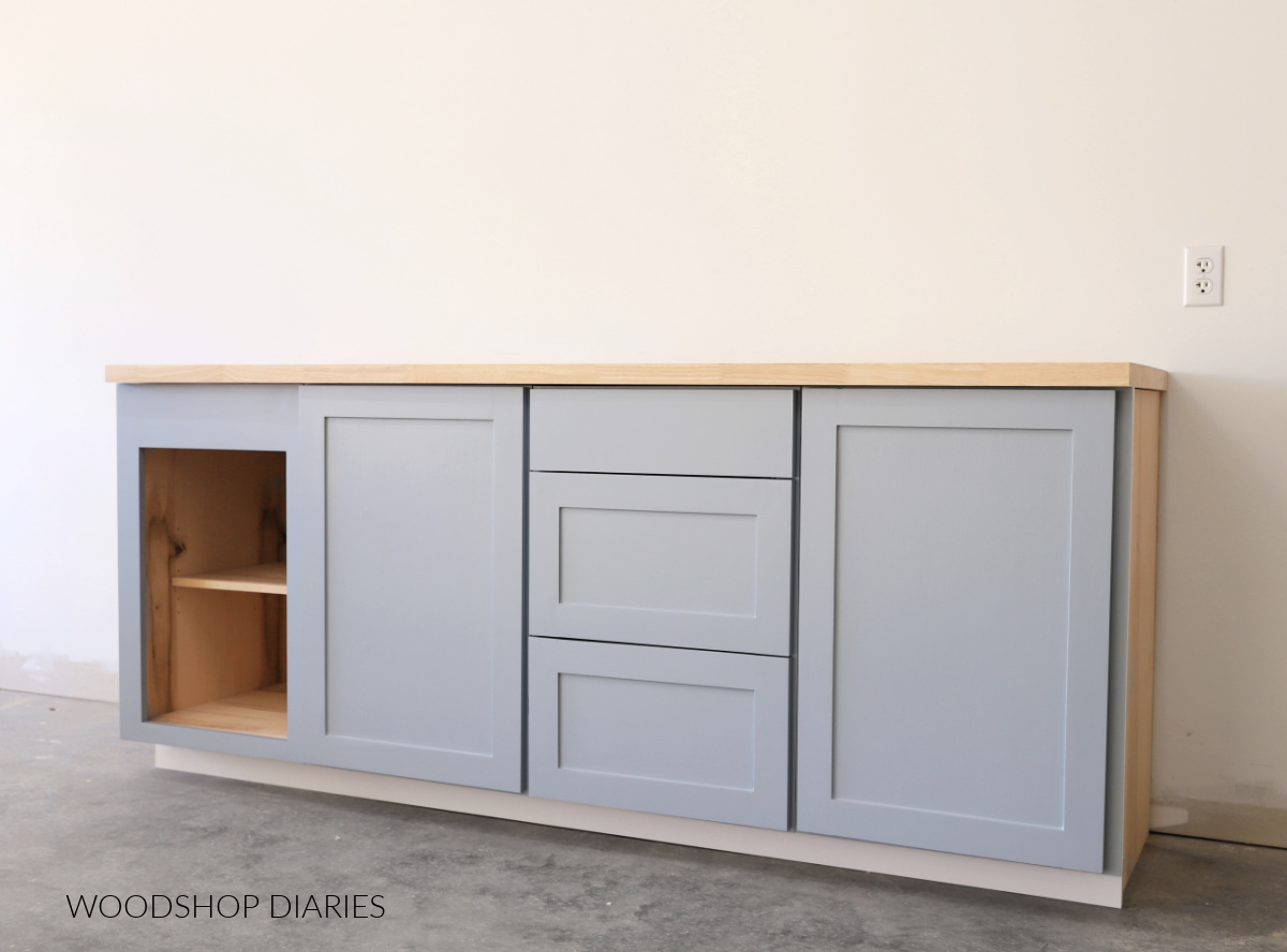 Simple basic base cabinets--one open, two with doors, and one with drawers--against white wall with butcherblock countertop