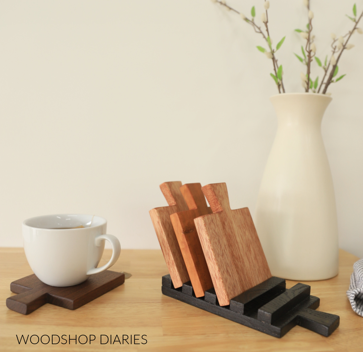three coasters placed in coaster tray, one coaster sitting on table top with cup of tea on top, vase in background