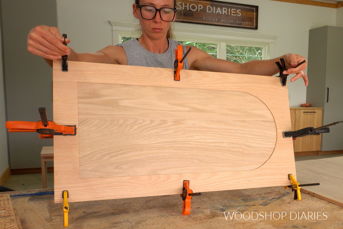 Shara Woodshop Diaries holding arched trim cabinet door with clamps on workbench