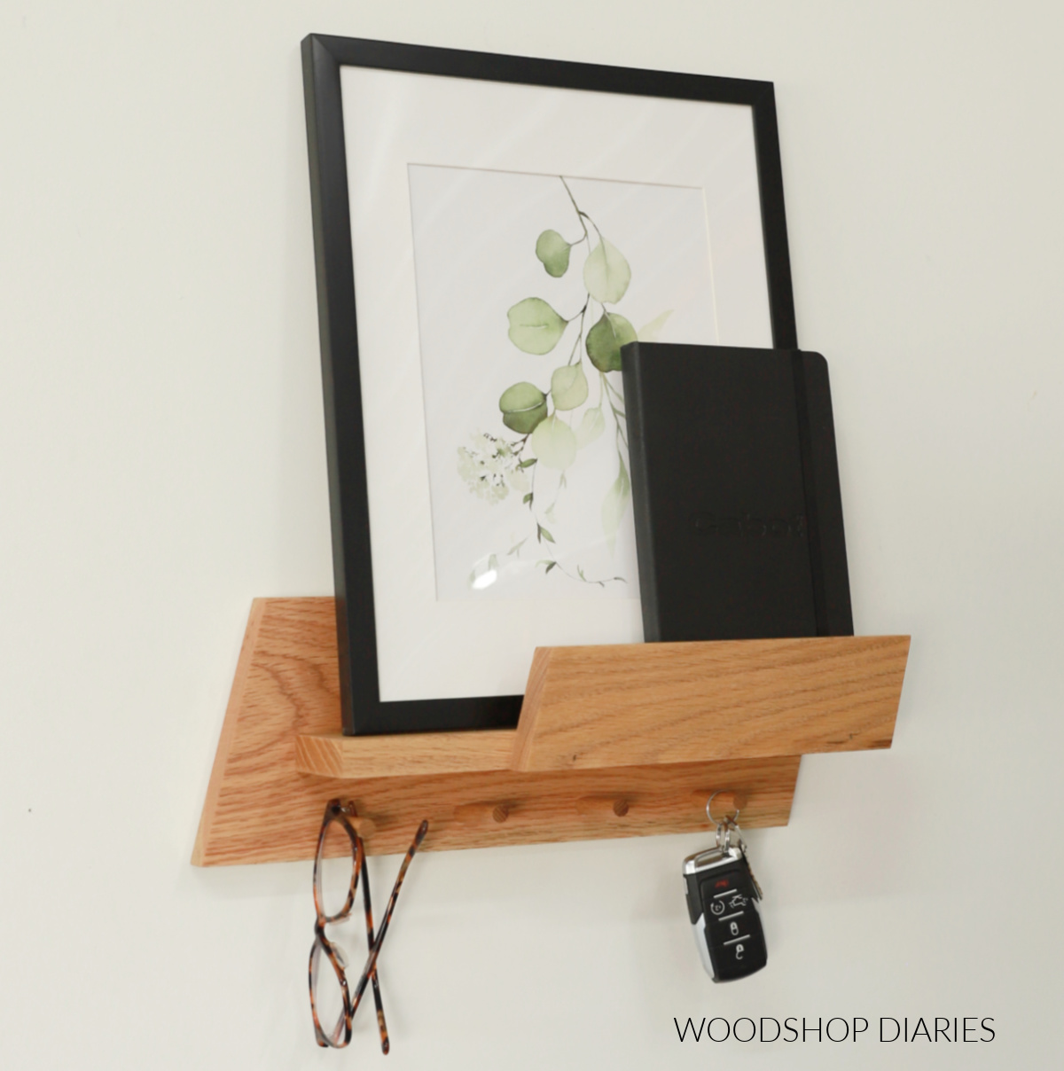 Completed wall shelf entryway organizer with picture and notebook setting on shelf and glasses and keys hanging on pegs