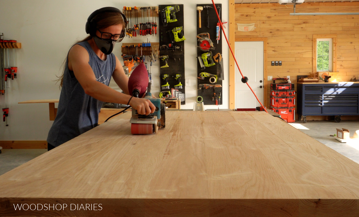 Shara Woodshop Diaries sanding table top with belt sander on workbench