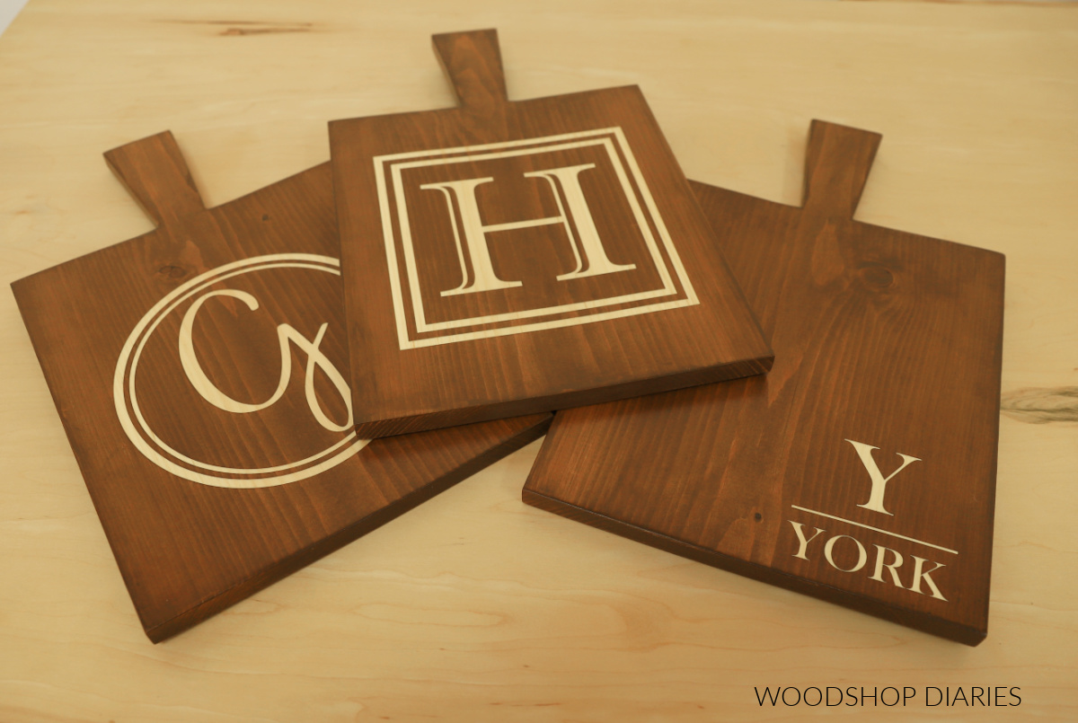 Three personalized serving boards stacked on workbench--one with round monogram, one with square monogram and one with last name in bottom corner