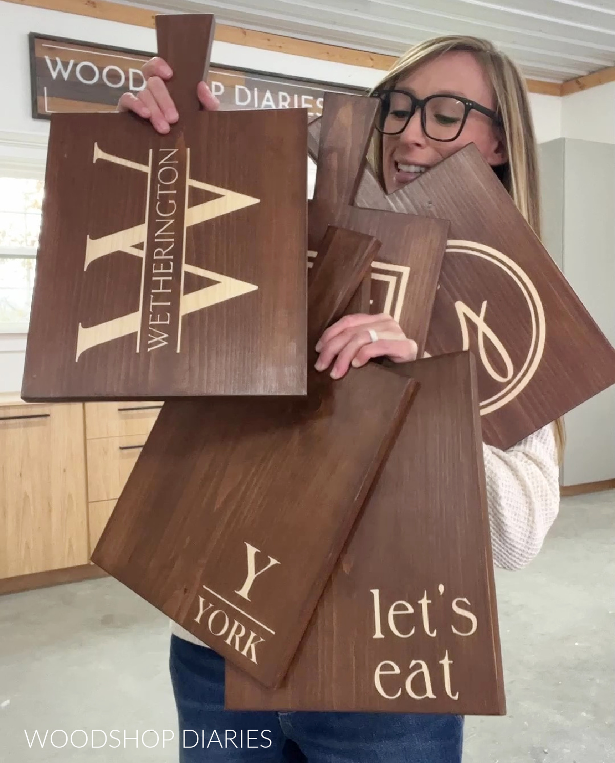 Shara Woodshop Diaries holding five customized serving boards with lettering stained on the front.